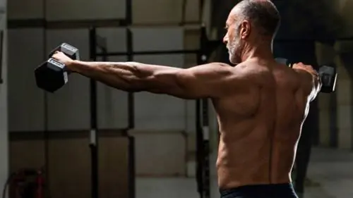 человек без рубашки в тренажерном зале, выполняющий упражнения на плечи с гантелями