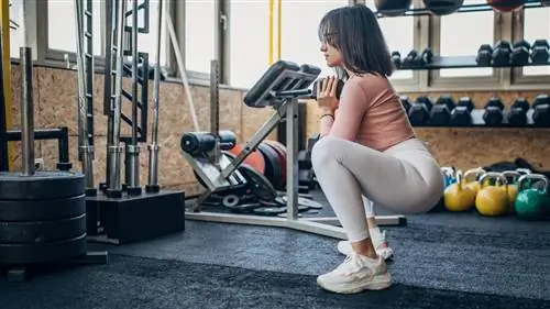 Женщина выполняет приседания с кубком, держа гантель