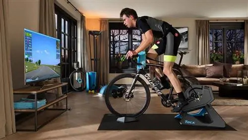 Мужчина едет на шоссейном велосипеде Specialized, прикрепленном к турботренажёру Tacx Neo 2T, перед телевизором с плоским экраном Мужчина едет на шоссейном велосипеде Specialized, прикрепленном к турботренажёру Tacx Neo 2T, перед телевизором с плоским экраном
