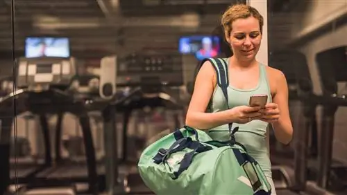 Женщина смотрит на свой телефон на фоне тренажерного зала, через плечо висит спортивная сумка, сгибание бицепса с гантелями Женщина смотрит на свой телефон на фоне тренажерного зала, через плечо висит спортивная сумка, сгибание бицепса с гантелями