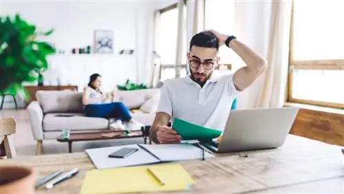 Мужчина потирает голову, глядя на документ. Он в гостиной, сидит за столом с открытым ноутбуком.